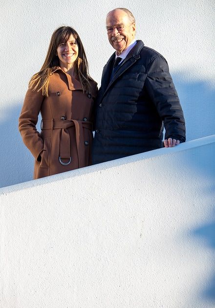 Mulher e homem de meia-idade, sorridentes, posam lado a lado ao ar livre, encostados a uma parede branca iluminada pelo sol; ela usa sobretudo castanho com cinto e tem cabelo comprido castanho, ele usa casaco acolchoado azul-escuro sobre camisa e gravata.